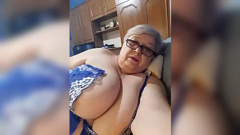 Media: Video of an older, large-breasted woman with short gray hair, wearing glasses and a blue lace bra, lying on a wooden bed in a dimly lit room.