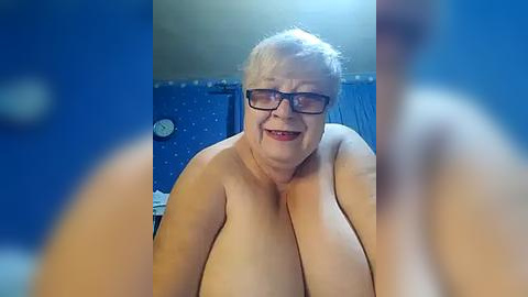 Media: Video of a smiling, older, overweight woman with short, gray hair and glasses, wearing glasses, topless, in a bathroom with blue walls, clock, and blurry background.