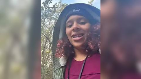 Media: A video of a young woman with curly hair, wearing a green hooded jacket and a maroon top, smiling outdoors in a forest with blurred trees in the background.