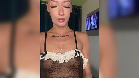 Media: A video of a light-skinned woman with short blonde hair, wearing a sheer black lace bra with white ruffles and a necklace, standing in a dimly lit room with a TV displaying a blurred image.