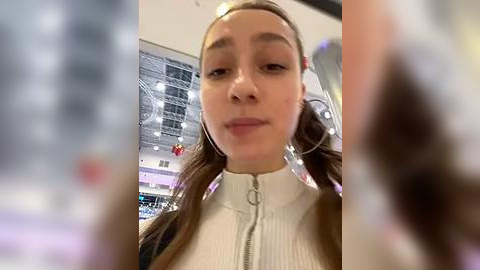 Media: A video of a young woman with fair skin, brown hair, and wearing a white ribbed sweater, taken indoors with blurred background featuring a digital display.