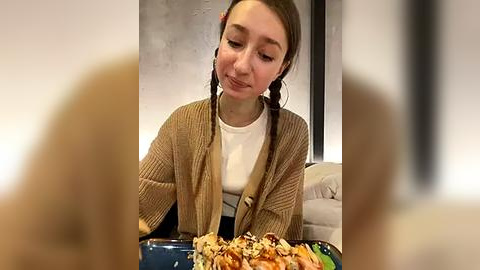 Media: Video of a young woman with light skin and long brown hair in pigtails, wearing a beige cardigan over a white shirt, eating a sandwich with nuts and seeds on a blue plate. The background shows a modern, minimalist room with a gray wall and a couch.