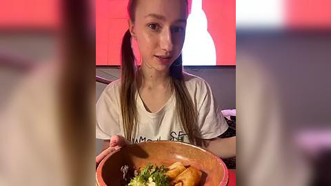 Media: Video of a young girl with light skin, long brown hair in pigtails, wearing a white T-shirt, holding a wooden bowl filled with salad, seated indoors under a red and white light backdrop.