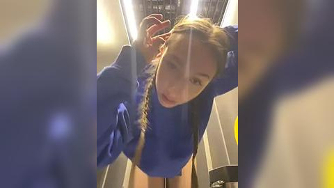 Media: Video of a young woman with fair skin and long brown hair, wearing a blue sweater, looking upwards in a dimly lit elevator, with blurred figures in the foreground.