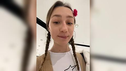 Media: Video of a young woman with long brown hair in braids, wearing a white shirt, beige cardigan, and a red flower in her hair, smiling, taken indoors with a blurred background.