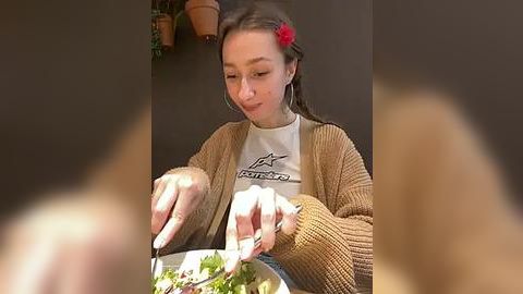 Media: Video of a young girl with light skin and long brown hair adorned with a red flower, wearing a beige knitted cardigan over a white graphic tee. She's eating salad in a restaurant setting with blurred out people in the foreground.