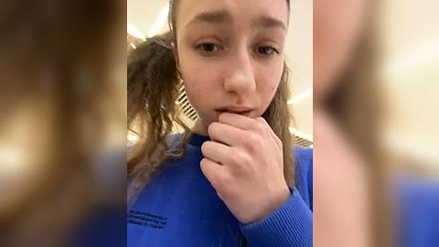 Media: Video of a young woman with fair skin, brown hair tied back, wearing a blue sweater, biting her finger thoughtfully, blurred background.