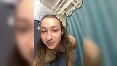 Media: Video of a young woman with straight, light brown hair in pigtails, wearing a beige sweater and blue jeans, smiling in a hospital room with teal curtains and white walls.