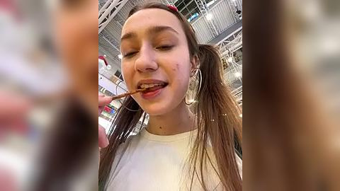 Media: Video of a young woman with long brown hair in pigtails, wearing large hoop earrings and a white shirt, licking an ice cream cone in a brightly lit, modern indoor space with industrial ceiling beams.