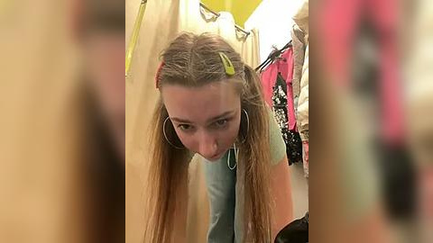 Media: A video of a young woman with long, blonde hair in pigtails, wearing a blue shirt and large hoop earrings, bending over in a cluttered, brightly-lit closet.