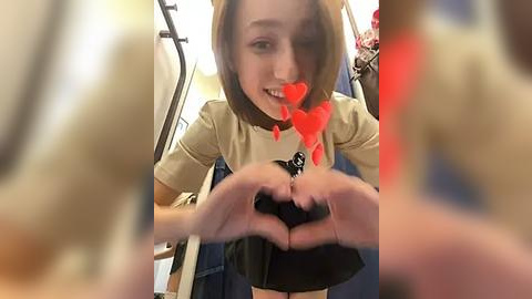 Media: Video of a young woman with shoulder-length hair, smiling and forming a heart shape with her hands, wearing a beige top and black skirt. Background shows a blurred indoor setting with a white curtain and red heart filters.