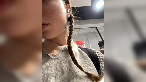 Media: Video of a young woman with light skin, brown braided hair, wearing a gray sweater, standing in a modern, dimly-lit room with a black backpack and red sign.