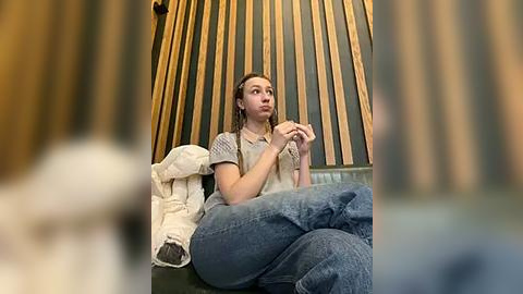 Media: Video of a woman with long, light brown hair in a gray t-shirt and jeans, sitting on a sofa with a beige blanket and a wooden cone-shaped lamp behind her.