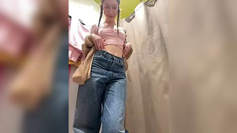 Media: Video of a young woman in a fitting room, adjusting her pink crop top and high-waisted jeans, with a beige handbag in hand, surrounded by beige changing room curtains.