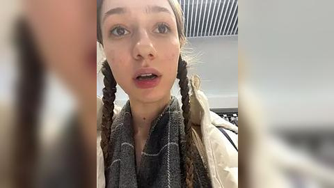 Media: Video of a young woman with light skin, brown hair in braids, wearing a gray scarf, looking surprised in a modern indoor setting with white walls and a ceiling.