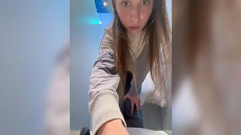 Media: A video shows a young woman with long brown hair, wearing a gray sweatshirt and gray pants, looking down while bending over a table in a dimly lit room.