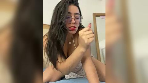 Media: Video of a Latina woman with long black hair, wearing glasses and a white lace dress, sitting on a bed, touching her feet, with blurred figures in the foreground.