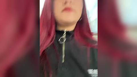 Media: Video of a woman with long, vibrant red hair and pale skin, wearing a black high-necked garment with a silver zipper, partially obscured by blurred red hair in the foreground.