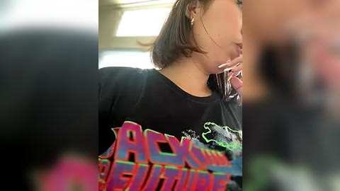 Media: Video of a young woman with shoulder-length brown hair, wearing a black t-shirt with \"Roxanne Revere\" in bold, colorful text, talking on a phone. Blurred background suggests a casual indoor setting.