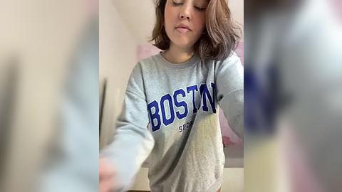 Media: Video of a young woman with shoulder-length brown hair, wearing a gray Boston College sweatshirt, taking a selfie in a dimly lit room with blurred objects in the foreground.