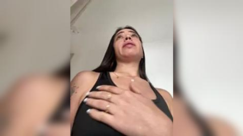 Media: A video of a young woman with long dark hair, wearing a black tank top, looking upward and covering her chest with her hands, in a minimalist white room with a ceiling fan.