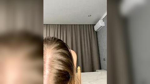 Media: A video showing a person with long, straight brown hair, facing away, in a modern, minimalistic bedroom with grey curtains, white ceiling, and air conditioner.