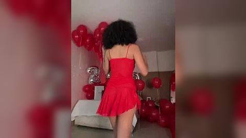 Media: Video of a woman with short, curly black hair wearing a red, sleeveless dress with a ruffled hem, surrounded by red balloons and a silver disco ball, standing in a brightly lit room.