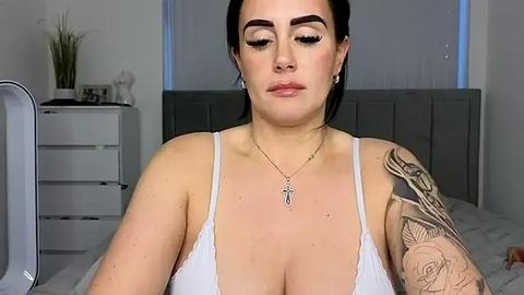 Media: A video of a fair-skinned woman with dark hair, wearing a white bralette, standing in a bedroom with a grey upholstered headboard, a white dresser, and a mirror. She has a tattoo sleeve and a cross necklace.