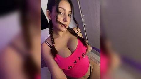 Media: Video of a Latina woman with medium skin tone and long black braided hair, wearing a pink tank top and beige underwear, gagged with tape, sitting in a dimly lit room.