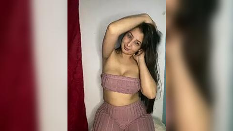 Media: Video of a young woman with long black hair, medium skin tone, wearing a strapless, pink, textured crop top and matching high-waisted skirt, posing against a white wall with a red curtain to the left.