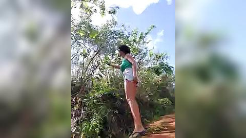 Media: Video of a woman in a green top and white shorts standing in lush, green foliage on a red dirt path under a bright blue sky.