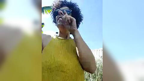 Media: A video captures a person with curly hair and sunglasses, wearing a yellow sleeveless dress, gesturing with their right hand, against a bright blue sky and green foliage.