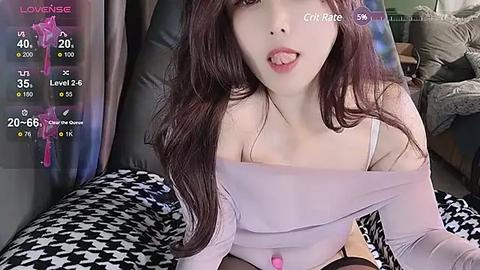 Media: Video of a young Asian woman with long, dark brown hair and fair skin, wearing a light purple off-shoulder top, lying on a black-and-white patterned bed. Background shows a cozy indoor setting with a TV screen displaying her \"Lovense\" stats.
