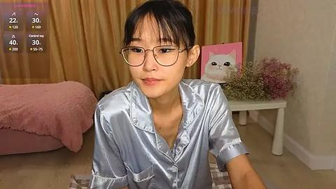 Media: Video of a young Asian woman with short black hair, wearing round glasses, a shiny blue satin shirt, and a pink blanket, in a cozy, softly lit bedroom with a white table and a framed cat painting.