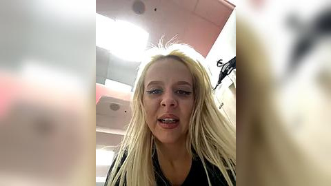 Media: A close-up video of a young woman with long blonde hair, wearing a black shirt, captured from a low angle. The background shows a blurred, pink ceiling.