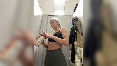 Media: Video of a blonde woman in a sports bra and high-waisted leggings, holding a medication bottle in a locker room with hanging coats and fluorescent lighting.