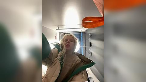 Media: Video of a blonde woman in a beige jacket, holding a green bag, in a laundry room with white walls and orange drying rack, blurry background.