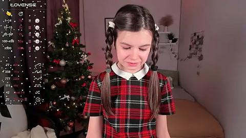 Media: Video of a young girl with long brown hair in pigtails, wearing a red and black plaid dress, standing in front of a decorated Christmas tree in a cozy, warmly lit room.