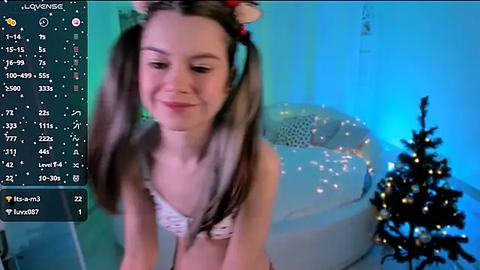 Media: A young, smiling woman with light skin, long brown hair in pigtails, wearing a white lace bra, stands in a cozy, lit room with a Christmas tree and fairy lights.