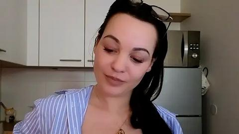 Media: Video of a fair-skinned woman with long dark hair, wearing a light blue striped shirt, and sunglasses on her head, standing in a modern kitchen with white cabinets and stainless steel appliances.