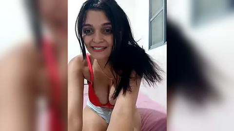 Media: Video of a smiling Latina woman with dark hair, wearing a red bra and gray underwear, leaning forward on a bed with pink sheets in a brightly lit room.