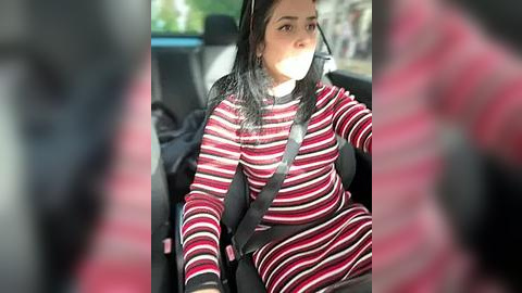 Media: A video of a woman wearing a striped red and black sweater, driving in a car with a seatbelt on. The background is blurred, showing a blurry, green park.