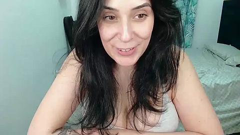 Media: Video of a middle-aged woman with long, wavy black hair, wearing a light-colored spaghetti-strap top, smiling, in a bedroom with a bed and teal curtains in the background.