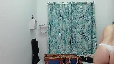 Media: Video of a bedroom with a woman in white polka-dot panties and a man's back, both facing away from the camera. The room has white walls, a green leaf-patterned curtain, and a white shelf with a white bird statue.