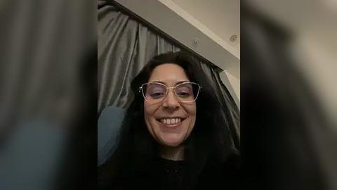 Media: Video of a woman with long black hair and glasses, smiling, lying on a gray bed with a white headboard, wearing a black top.