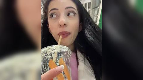 Media: Video of a young woman with long black hair and fair skin, drinking a green smoothie through a straw, wearing a white shirt, indoors, blurred background.