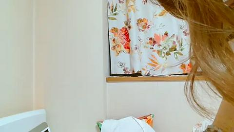 Media: Video of a small, bright bathroom with a floral-patterned shower curtain, beige walls, a white sink, and a wooden window sill. A person's shoulder and hair are partially visible in the foreground.