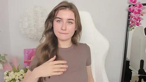 Media: Video of a young woman with long, wavy brown hair, wearing a light brown top, seated in a white chair, surrounded by white and pink floral decor.