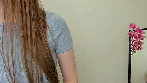 Media: Video of a woman with long, straight, light brown hair, wearing a light blue ribbed t-shirt, standing against a plain cream-colored wall. A floral arrangement with pink flowers is visible in the background.