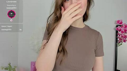 Media: Video of a young woman with fair skin, long brown hair, wearing a ribbed, short-sleeve, brown top, covering her mouth with her hand. Background features a pink flower arrangement and a digital overlay with social media icons.